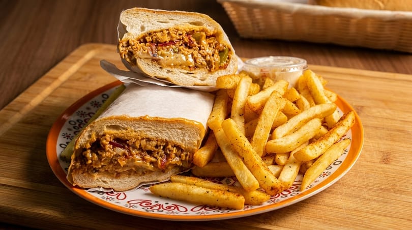 A chicken cheesesteak sandwich, cut in half, with a side of seasoned french fries and dipping sauce.