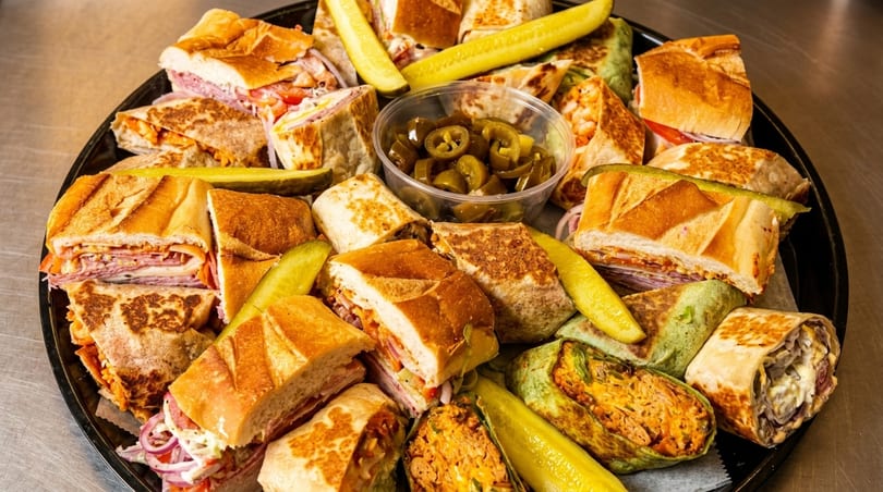 A catering platter filled with various sandwiches, wraps, pickles, and a container of sliced jalapeños.