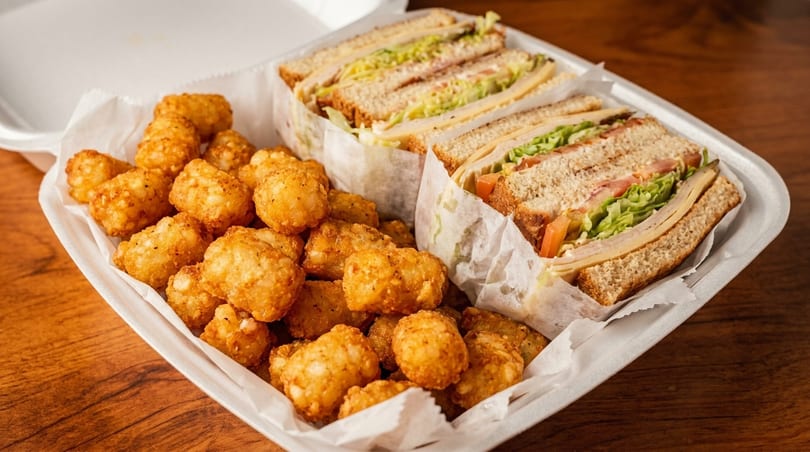 A white takeout container holds a club sandwich cut into halves and a pile of golden tater tots.