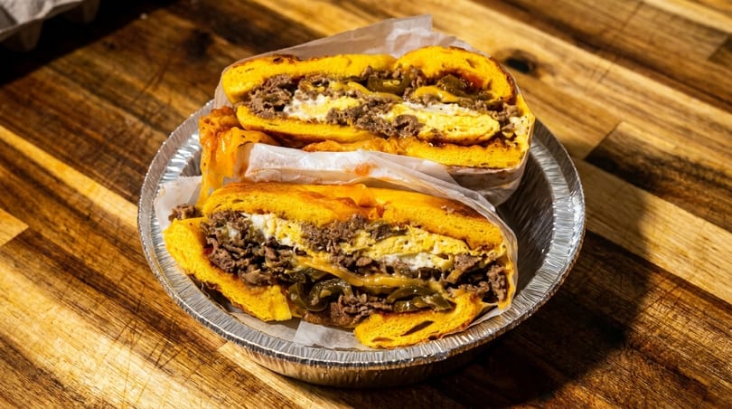 Two halves of a hearty breakfast sandwich with egg, meat, cheese, and peppers in a foil container.