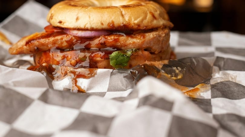 Close-up of a chicken sandwich with red onions and sauce on a bagel, wrapped in checkered paper.